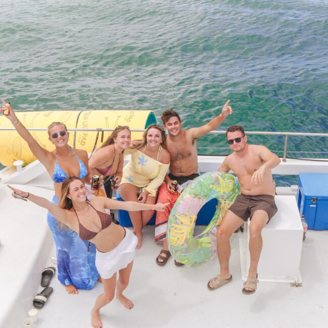 Six smiling people in swimsuits pose for a photo on a boat surrounded by blue ocean water, with two life rings, large inflatable floats, and coolers visible on deck.