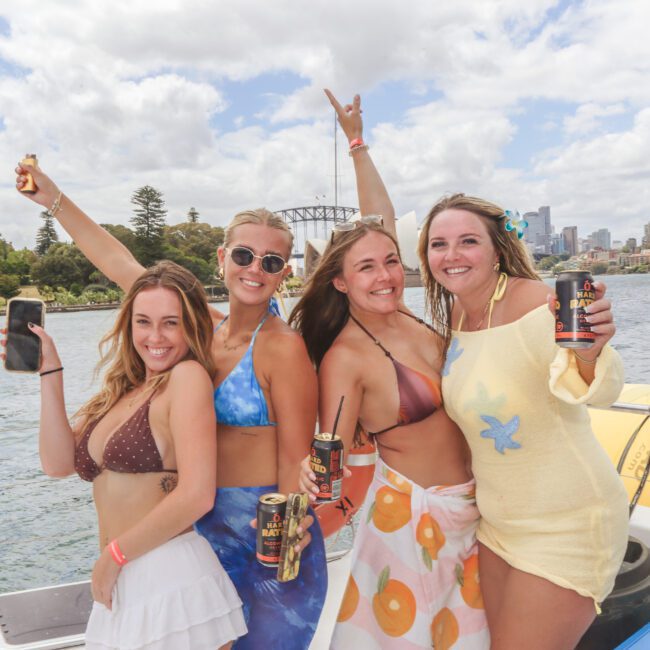 Four women in swimwear smile and pose with drinks on a boat, with water, greenery, and a cityscape, including a bridge, in the background under a partly cloudy sky.
