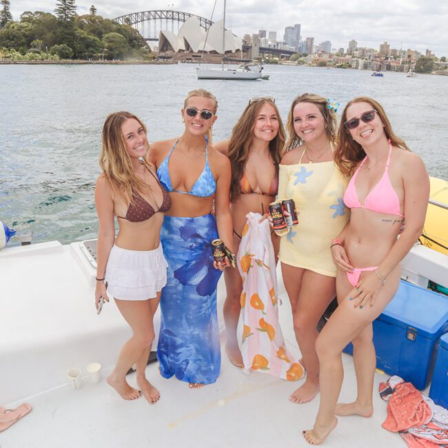 Five women in swimsuits smile and pose on a boat with drinks in hand. The Sydney Opera House and city skyline are visible in the background across the water on a cloudy day.