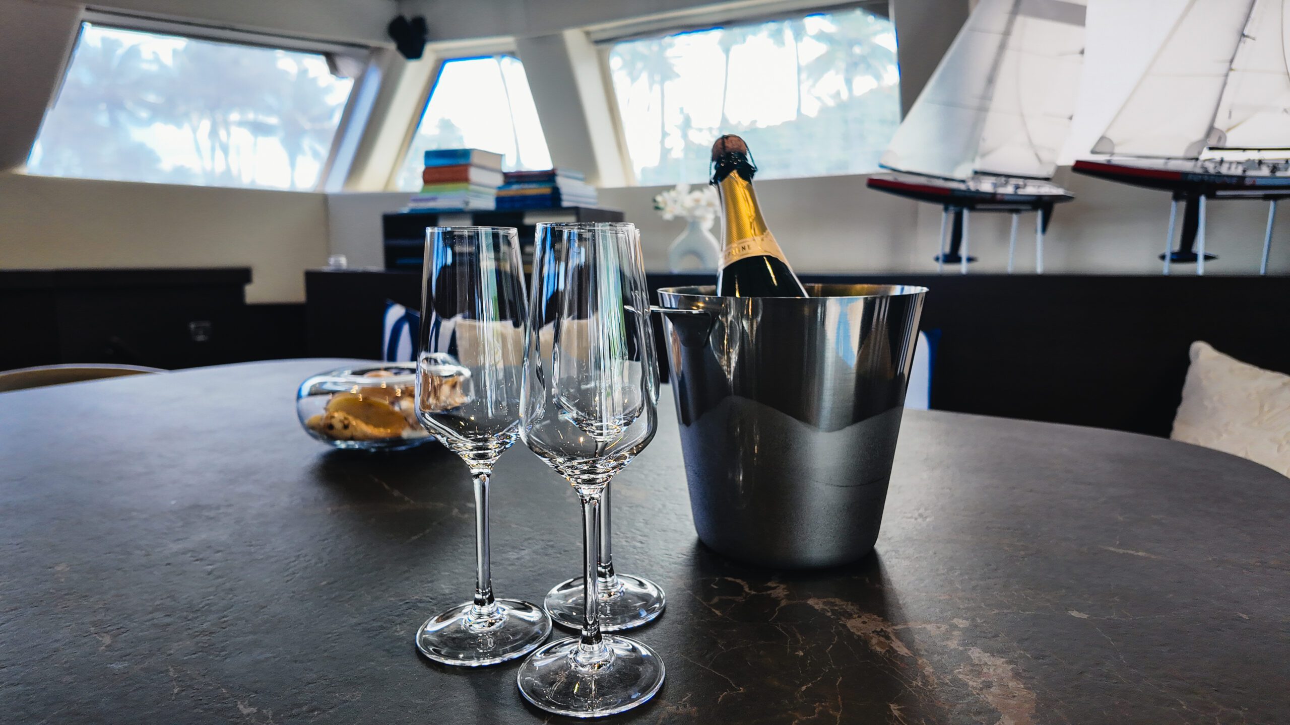 Three empty champagne glasses and a bottle of champagne in an ice bucket are on a table inside a room with large windows, model sailboats, books, and a bowl of snacks in the background.