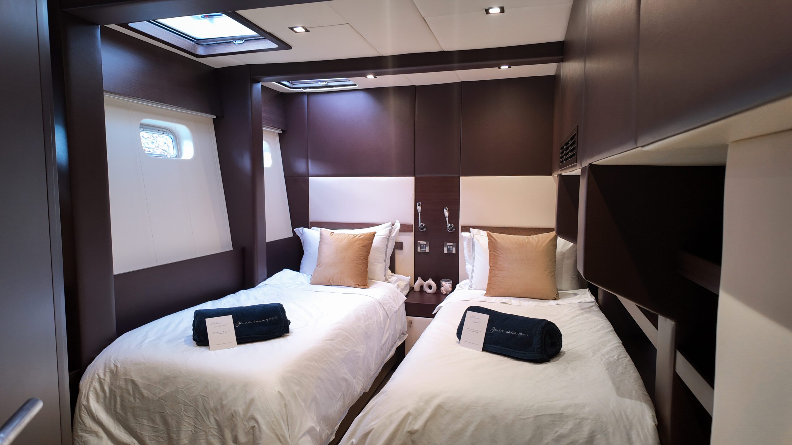 A modern cabin with two single beds, each topped with beige pillows and folded dark towels. The room has brown and white walls, built-in lighting, and a small window on the left.