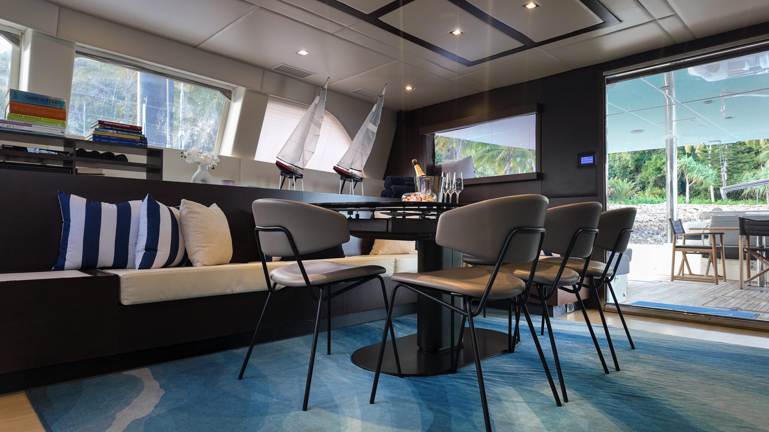 Modern yacht interior with a dining table, six chairs, and a built-in cushioned bench with striped pillows. Large windows provide natural light and views of greenery outside. A deck is visible through an open glass door.