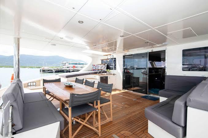 The SV Feng yacht deck features a wooden dining table with chairs, gray cushioned seating, and large glass doors that open to the interior, all overlooking a scenic waterfront view.