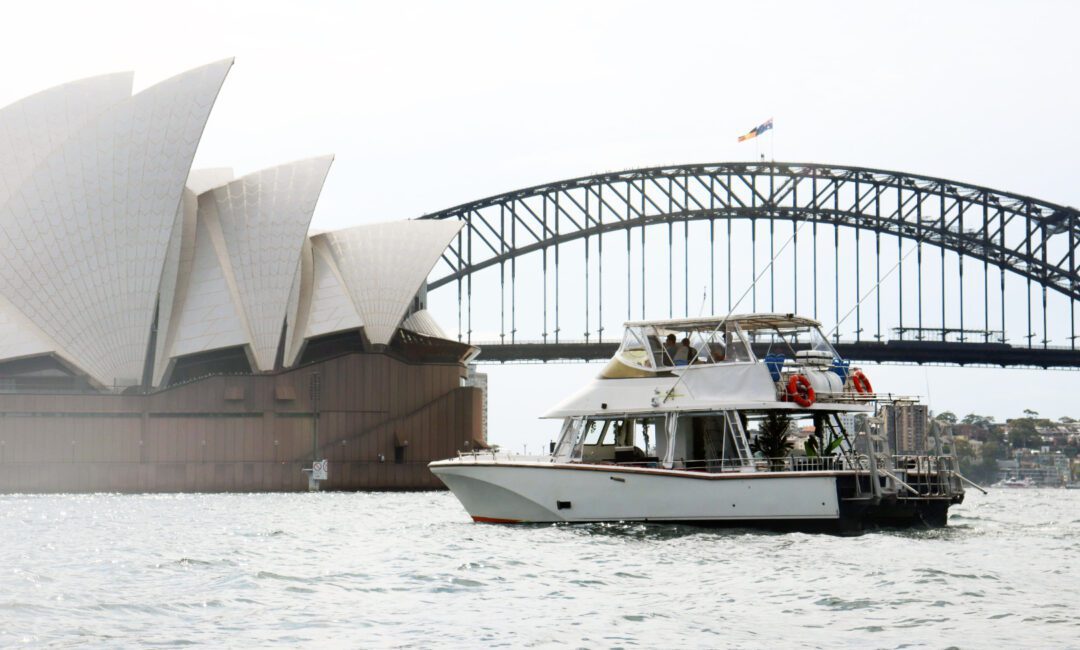 Curious George admires the iconic Sydney Opera House and Harbour Bridge from a white boat floating on Sydney Harbour on a cloudy day.