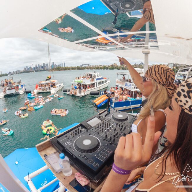 Two women in animal print outfits DJ on a boat overlooking a lively party on the water, with yachts, floats, and people swimming nearby. The city skyline is visible in the background under a cloudy sky.