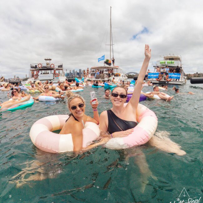 Two women in swimsuits smile and pose in pastel pool floats, holding drinks, surrounded by people on floats and boats at a lively water party. City skyline is visible in the background.