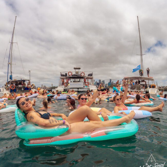 A group of people relax on colorful pool floats in the water near several anchored yachts under a cloudy sky, with a city skyline visible in the background. Some are smiling and raising their arms, enjoying the event.