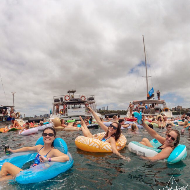 Three people relax on colorful pool floats in the water, smiling at the camera. Other people on floats and several boats are visible in the background under a cloudy sky. The scene is festive and social.