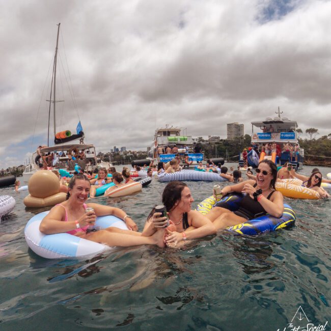 A group of people relax and laugh on inflatable pool floats in the water near docked boats, enjoying drinks under a cloudy sky. Buildings and more boats are visible in the background.