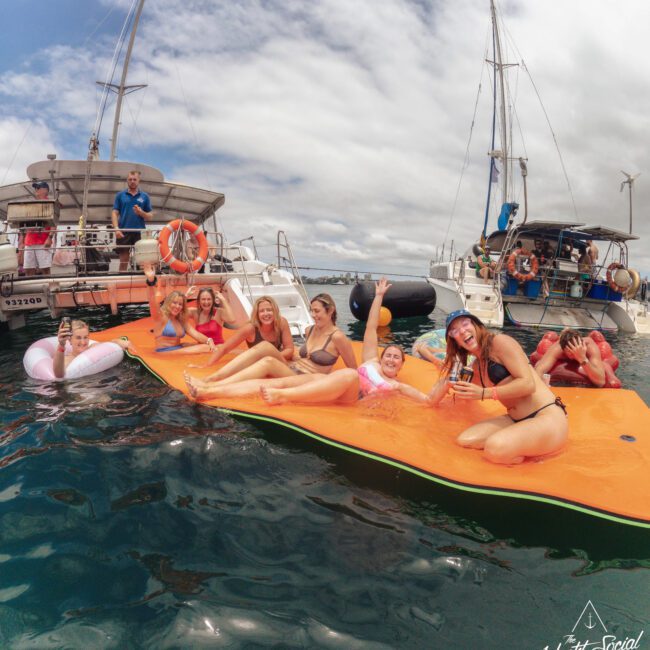 A group of people in swimsuits relax and pose on an orange floating mat in the ocean, with two yachts anchored nearby under a cloudy sky. Some are smiling and waving at the camera.