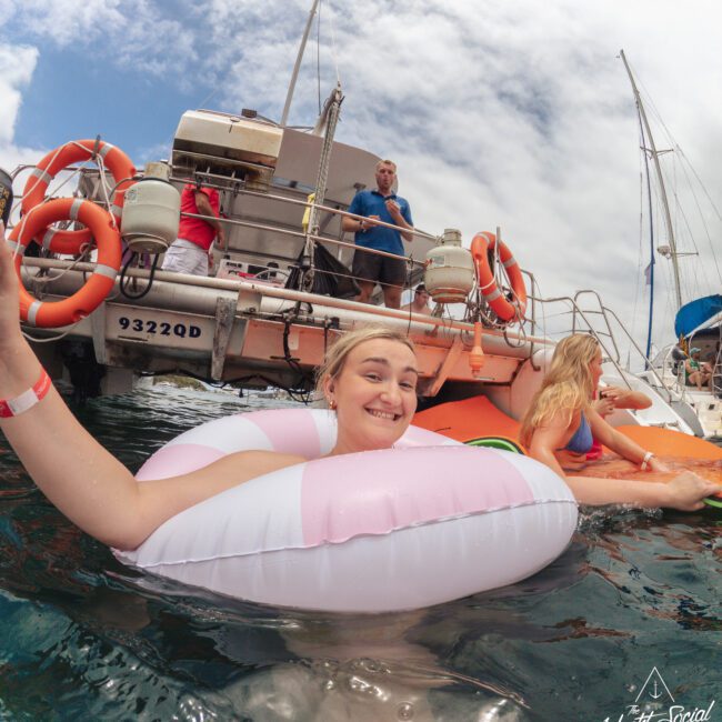A person smiling in a pink and white pool float in the ocean near a boat, with another person on an orange float beside them. Several people stand on the boat in the background under a partly cloudy sky.