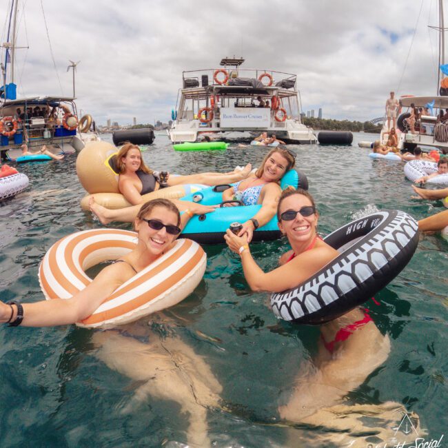 Four women in swimsuits float on colorful inflatable rings in the water, smiling and holding hands. Boats are anchored nearby under a partly cloudy sky. The scene is lively and joyful, suggesting a fun summer outing.