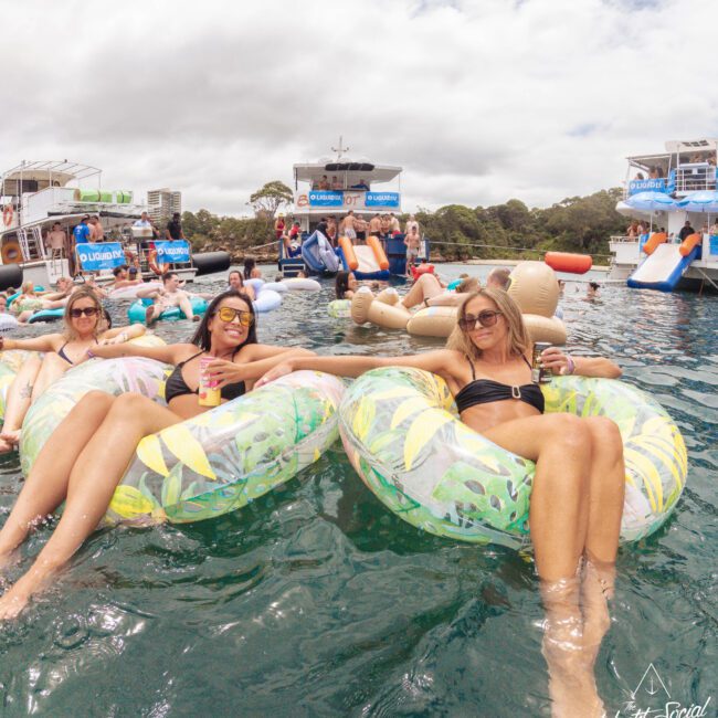A group of women relax on colorful pool floats in the water, smiling and enjoying drinks. Several boats with people partying are anchored in the background under a cloudy sky.