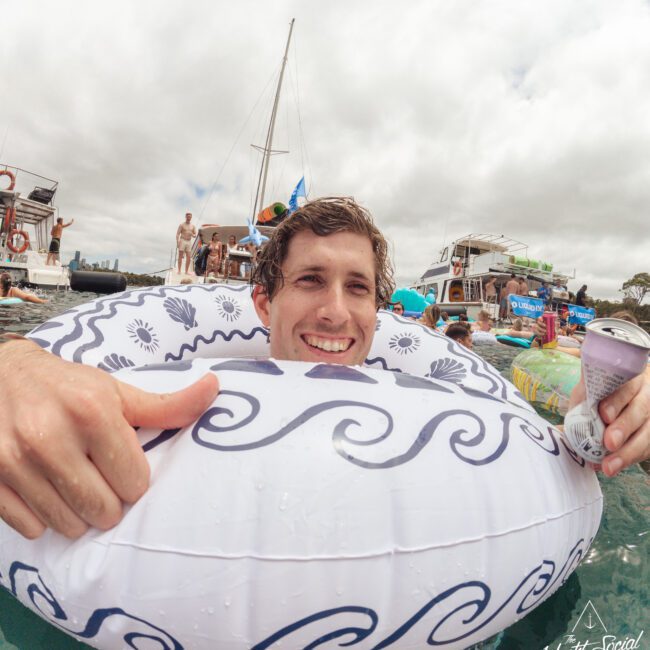 A smiling person in a patterned inflatable ring holds a drink and gives a thumbs up while floating in the water near boats and people on a cloudy day.