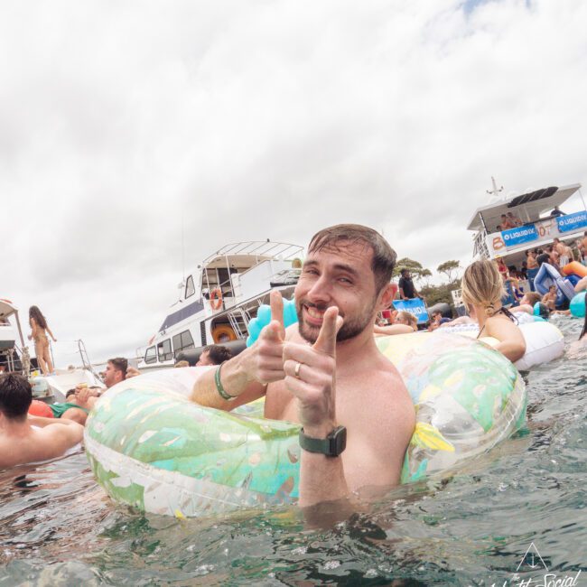 A smiling man in a pool float points both fingers at the camera, surrounded by people in the water near docked yachts on a cloudy day. The watermark reads “Yacht Social Club” in the bottom right corner.