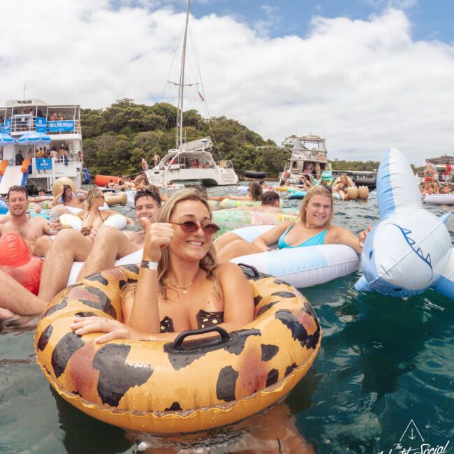 A group of smiling people relax on colorful pool floats in the water near anchored yachts, with trees and a partly cloudy sky in the background. A woman in leopard-print sunglasses sits in a leopard-print float.