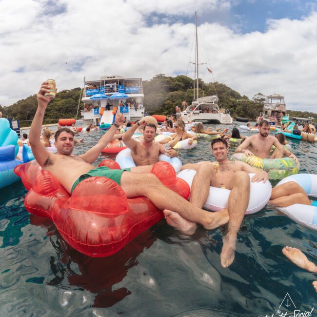 Five men relax on inflatable floats in the water, surrounded by other people on floats and boats. They are smiling, some holding drinks, with a backdrop of blue water, green trees, and partly cloudy skies.