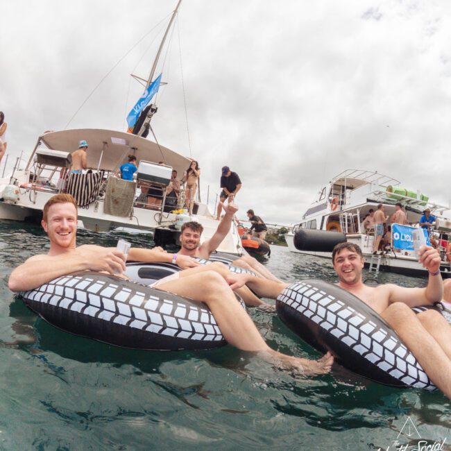 Three smiling men relax on inflatable tubes in the water, holding drinks. Behind them, people socialize on two nearby boats under a cloudy sky, enjoying a lively boating event. The water is calm and festive.