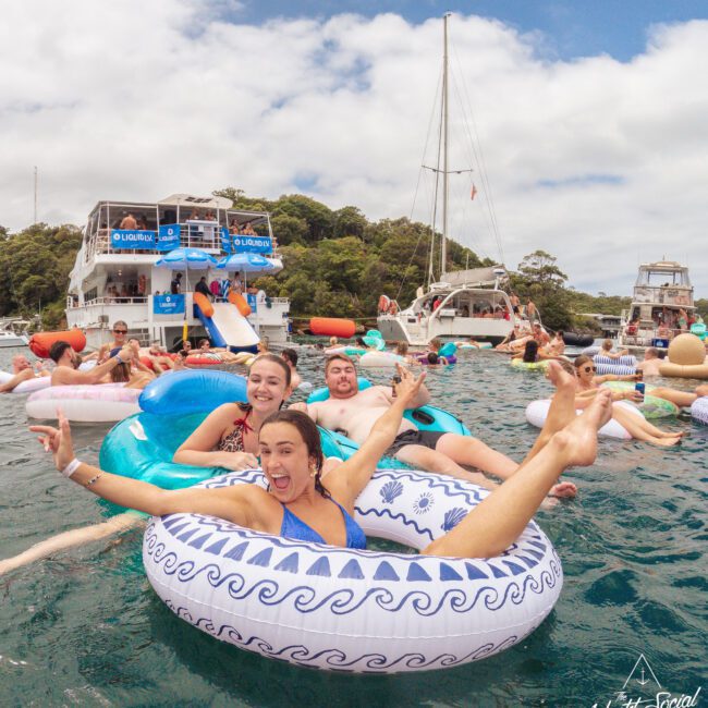 Three people smiling and posing on inflatable tubes in the water near boats, surrounded by others enjoying a sunny day at a lively floating party. The atmosphere is festive and relaxed.