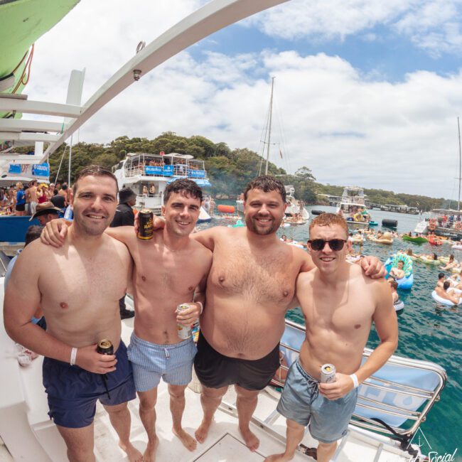 Four men in swim trunks stand arm-in-arm, smiling on a boat with drinks in hand. Behind them, people relax on boats and inflatables in a sunny, scenic bay with green hills and blue water.