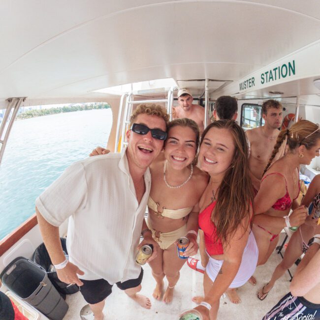 A group of young adults in swimwear smile and pose together on a boat, holding drinks, with water and trees visible outside the boat window. The mood is cheerful and relaxed.