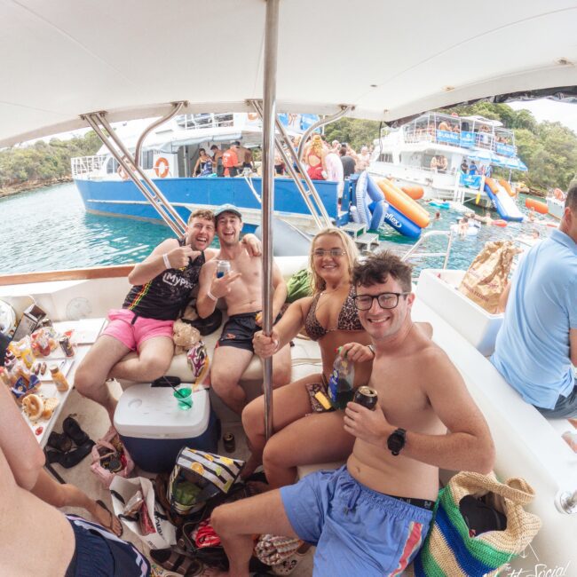 Four young adults in swimsuits sit smiling on a boat, holding drinks. The boat is surrounded by water, with more people and boats, including a large blue one, visible in the background on a sunny day.