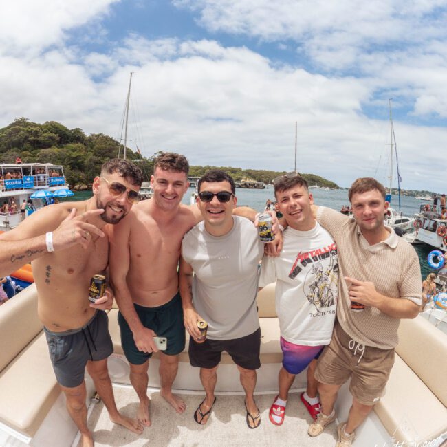 Five young men stand close together on a boat, smiling at the camera and holding drinks. Boats and water are visible in the background under a partly cloudy sky.