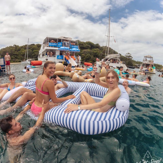 A group of people relax on inflatable floats in the water near anchored boats, socializing and enjoying drinks under a partly cloudy sky. Trees line the shore in the background.