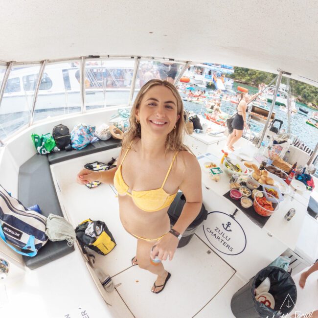 A smiling woman in a yellow bikini stands on a boat with food, drinks, and bags around her. People and colorful kayaks can be seen on the water and dock in the background. The scene is bright and lively.