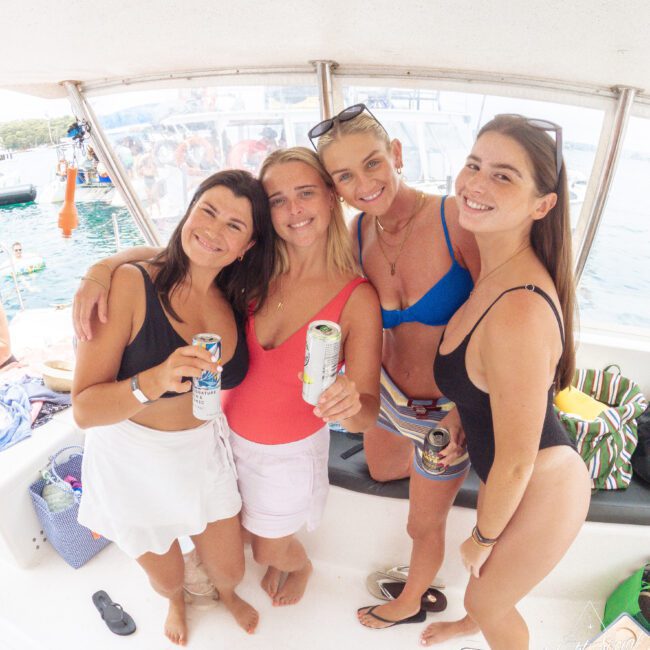 Four women in swimsuits smile and pose together on a boat, each holding a canned drink. The boat is docked on clear blue water, with a group of people visible in the background. It’s a bright, sunny day.