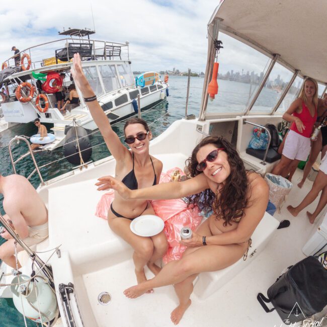 Two women in swimsuits sit on a boat, smiling and waving at the camera. Other people and boats are visible in the background on the water. The sky is partly cloudy, creating a cheerful and relaxed atmosphere.