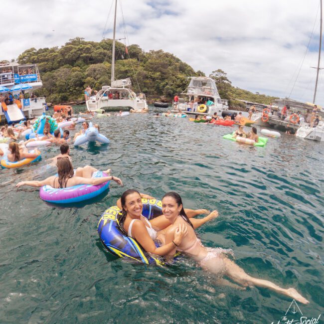 Two women smile and pose on an inflatable tube in the water, surrounded by others on floats and boats. It’s a lively scene with people enjoying a sunny day near anchored yachts. "Yacht Social Club" logo is in the corner.
