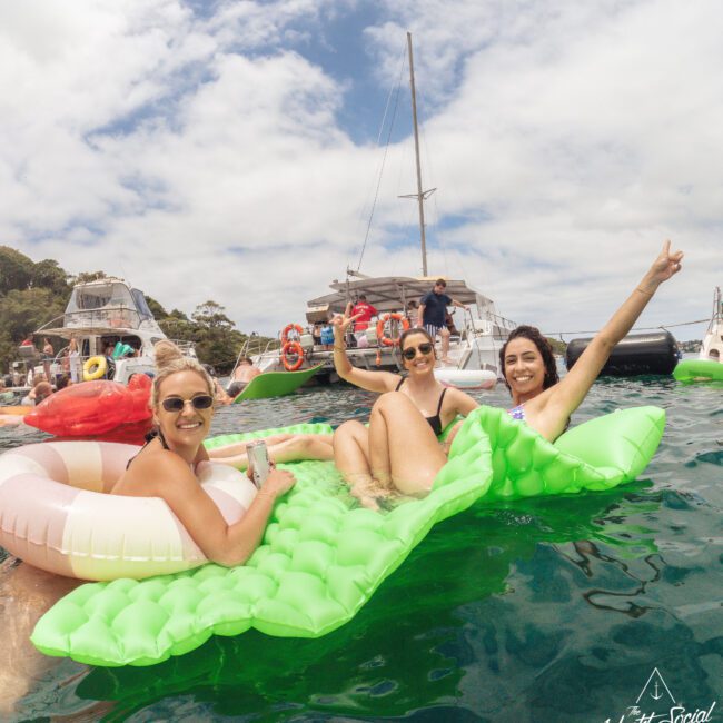 Three smiling women relax on inflatable pool floats in the water near anchored boats, enjoying a sunny day. Other people and boats are visible in the background under a partly cloudy sky.