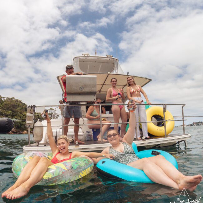 Two women relax on inflatable tubes in the water, smiling and raising drinks. Behind them, five people stand and sit on the deck of a boat, some holding drinks. The scene is lively and sunny, with water and trees in the background.
