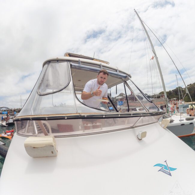 A man leans out from the window of a white boat, giving a thumbs-up gesture. Other boats and people are visible in the background on the water under a cloudy sky.