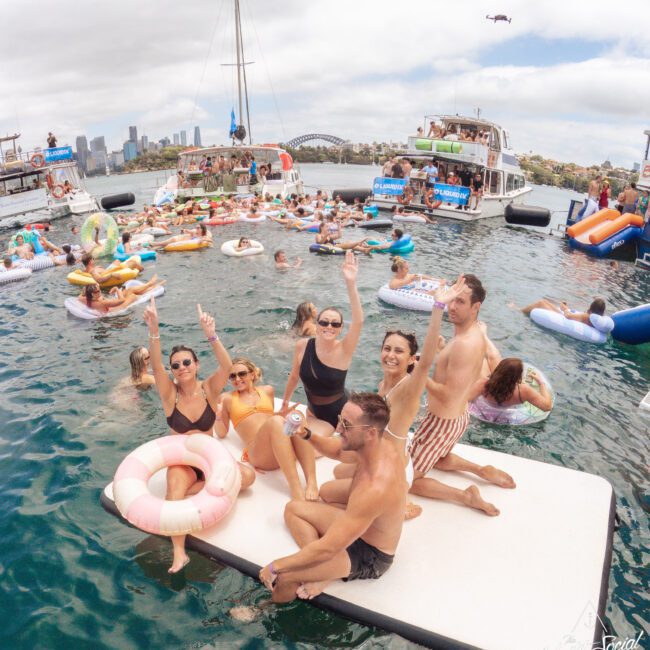A group of people in swimsuits relax and pose on a floating mat in the water, surrounded by others on inflatables and boats. The scene is lively, with city buildings and a bridge visible in the background.