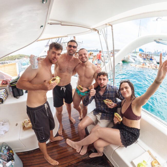 Five people in swimwear smile and pose for a photo on a boat, holding food and drinks. The boat is on clear blue water, with other boats and people visible in the background on a sunny day.