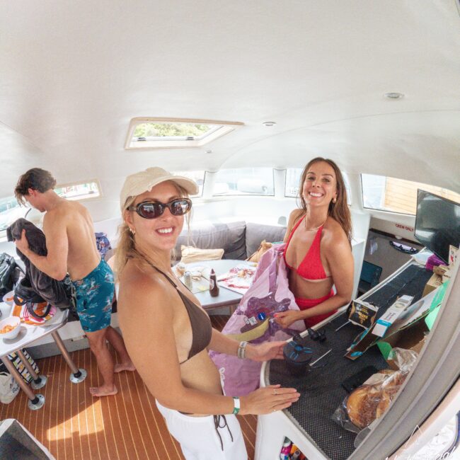 Two women in swimsuits and sunglasses smile at the camera inside a boat cabin; a man in swim trunks stands nearby. The table and counter have drinks, snacks, and bags, suggesting a relaxed, social atmosphere.