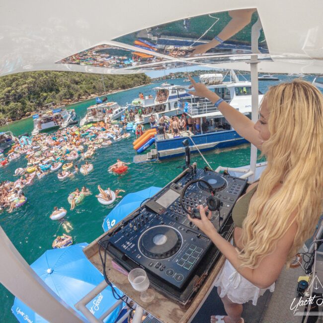 A DJ with long blonde hair plays music on a yacht, overlooking a crowd of people partying and floating on inflatables in bright blue water, surrounded by boats and lush green trees.