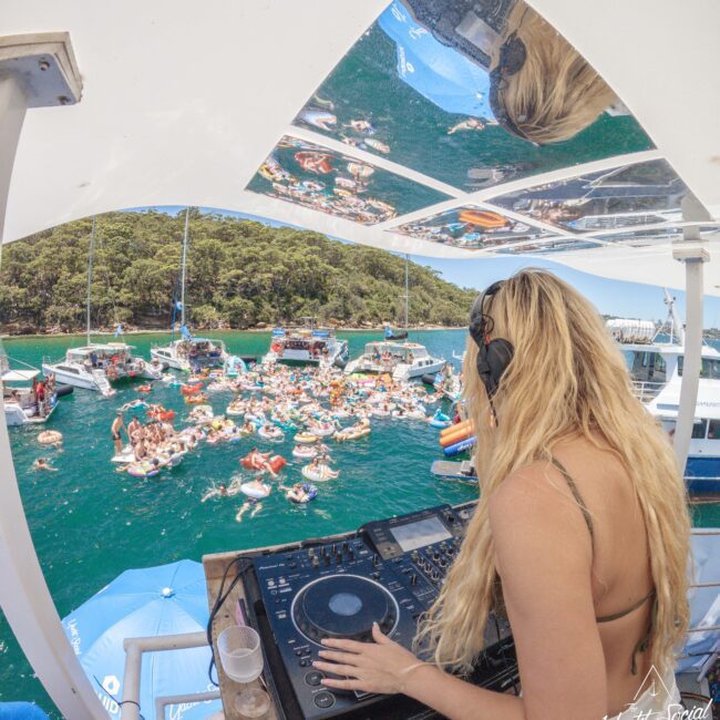 A blonde DJ wearing headphones plays music on a boat deck overlooking a crowd of people floating on inflatables in turquoise water, with boats and forested hills in the background.