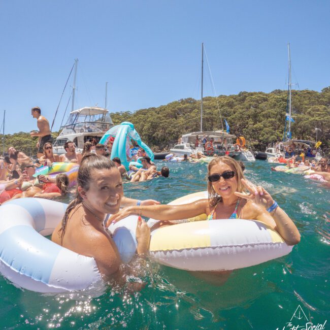 Two smiling women in inflatable pool rings float in clear blue water at a lively boat party, surrounded by other people, boats, and lush green trees under a sunny sky.