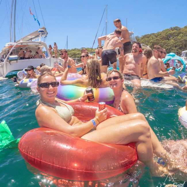 A group of people enjoy a sunny day on the water, lounging on colorful inflatables and swimming near boats. Two women in sunglasses smile on a red float, holding drinks, surrounded by other partygoers in swimsuits.