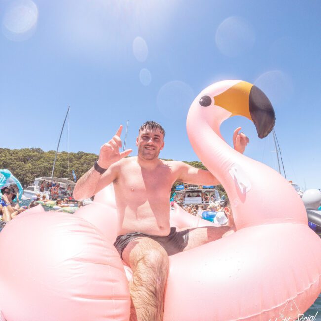 A smiling man in swim trunks sits on a large pink inflatable flamingo in the water, surrounded by boats and other floaties, under a bright blue sky.