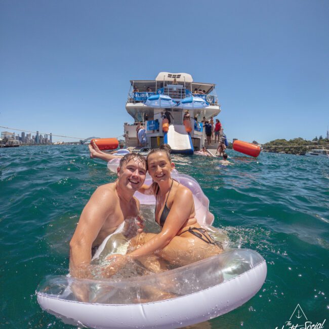 A smiling couple in swimsuits sits on a pool float in the water near a large yacht, with people visible on the boat and city buildings in the distant background under a clear blue sky.
