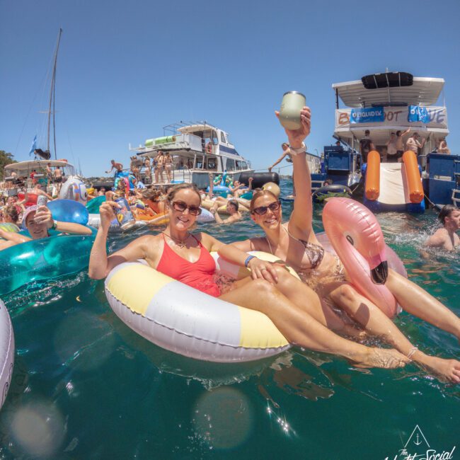 Two smiling women in swimsuits float on inflatable pool rings, one shaped like a flamingo, raising drinks. They are surrounded by other people enjoying a lively boat party on clear blue water under a sunny sky.