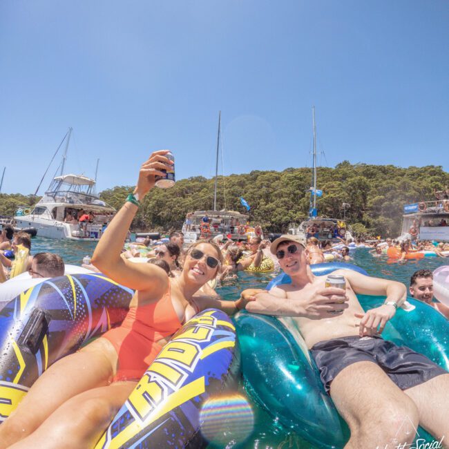 Two people relax on colorful inflatable floats in the water, smiling and holding drinks. Behind them, many others enjoy the sunny day with boats and lush green trees in the background.
