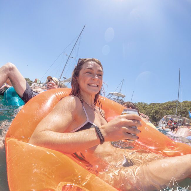 A smiling woman in a bikini holds a drink while relaxing on an orange inflatable float in the water, with boats and people in the background under a clear blue sky.