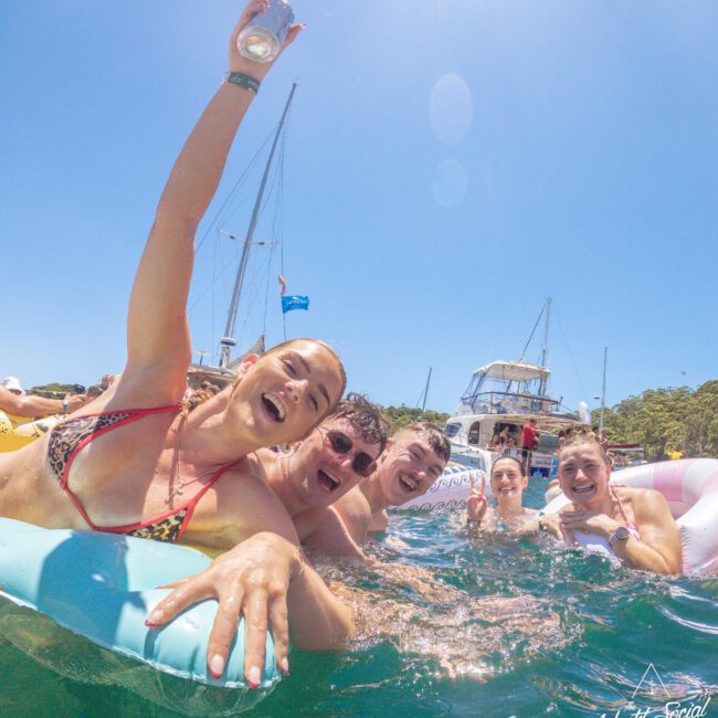 A group of young adults laugh and pose together while swimming and floating on inflatables in clear blue water, with boats and a sunny sky in the background. One person raises a drink, celebrating.