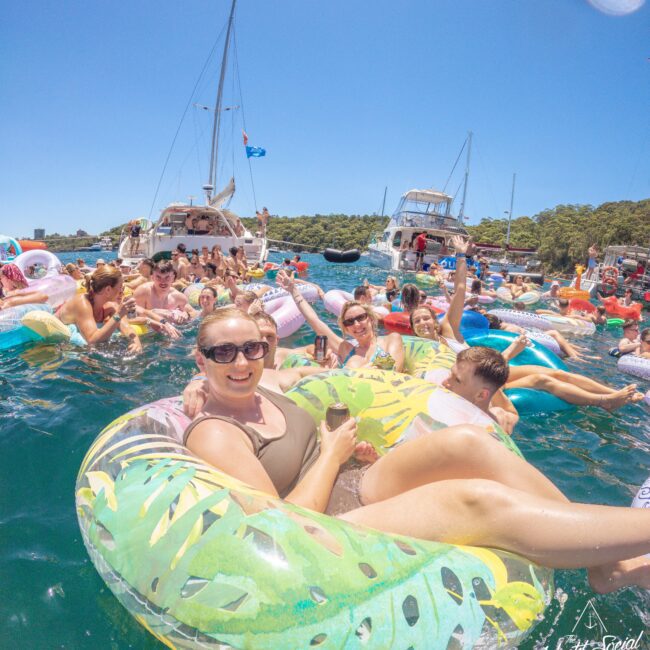 A group of people relax on colorful inflatable pool floats in the water, smiling and holding drinks, with boats and lush green trees in the background under a clear blue sky.