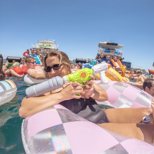 A person in sunglasses lounges on a pink and gray inflatable in the water, holding a colorful water gun. There are other people on inflatables and boats in the background, enjoying a sunny day.